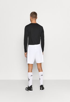 Mężczyzna stojący tyłem, ubrany w czarną koszulkę z długim rękawem, białe spodenki piłkarskie, białe skarpety Adidas i czarne korki, stojący przy jednolitej ścianie.