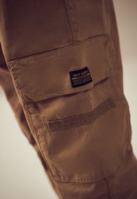 Poche de pantalon cargo marron avec rabat et étiquette noire portant la mention « Next Supply » cousue sur le côté de la jambe.