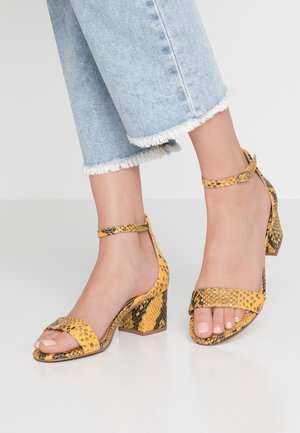 Füße in gelben Sandalen mit Schlangenmuster, offenem Zehenbereich und Blockabsatz, kombiniert mit hellblauen, ausgefransten, gekürzten Jeans auf neutralem Hintergrund.