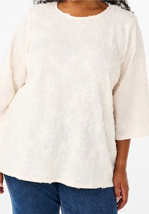 Jasnokremowy, teksturowany top z kwiatowym wzorem, prążkowaną tkaniną, okrągłym dekoltem i rękawami do łokcia, w zestawieniu z dżinsami.