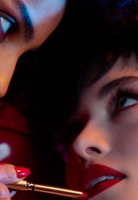 Close-up of two faces; one applying red lipstick using a gold applicator. Smooth skin textures, prominent lashes, colorful lighting effects.