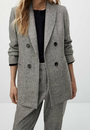 Woman wearing a grey checkered double-breasted blazer and matching trousers with a black top underneath, standing against a plain background.