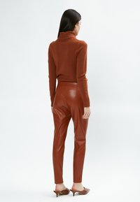 Maglione a collo alto in rust con polsini a coste, abbinato a pantaloni attillati in finta pelle lucida marrone e mule aperte marroni.