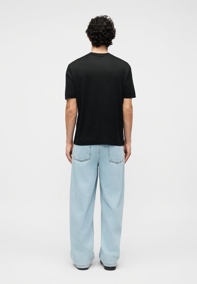 Άνδρας με σγουρά μαλλιά, φοράει μαύρο πουκάμισο με κοντά μανίκια και ανοιχτόχρωμα τζιν με φαρδύ πατζάμι, στέκεται με την πλάτη προς τα εμπρός σε απλό φόντο.