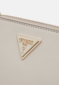 Cremefarvet tekstureret stofpung med et guldtonet trekantet logo, der viser "GUESS" og et spørgsmålstegn, samt lynlås lukning.