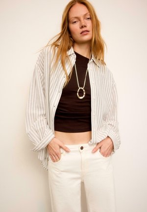 Žena s dlhými červenými vlasmi, oblečená v béžovej pruhovanej košeli prekrytej čiernym skráteným topom, biele nohavice a dlhý zlatý oválny prívesok na náhrdelníku.