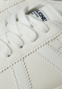 Weiße synthetische Sneaker mit strukturiertem Material, Schnürverschluss und Zickzacknähten. Markenlogo sichtbar auf dem Zungenlabel.