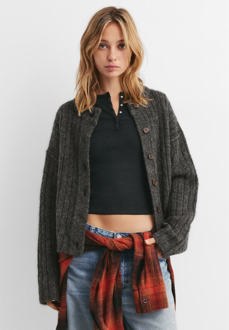 Cardigan a coste di un grigio scuro con chiusure a bottone, indossato sopra un top nero attillato e completato da una camicia a quadri rossa legata in vita.