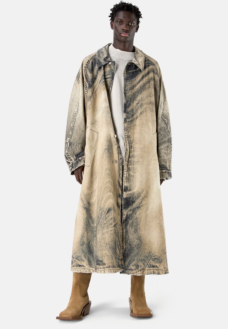 Homme noir debout, portant un long manteau beige et noir à motifs sur un pull blanc avec des bottes en daim beige.