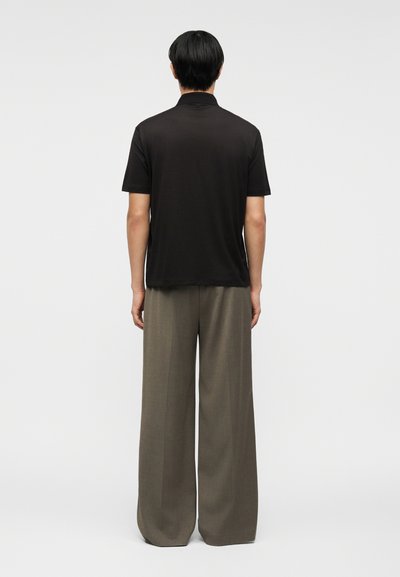 Άτομο που στέκεται με την πλάτη γυρισμένη, φοράει μαύρο κοντομάνικο πουκάμισο και άνετο γκρι παντελόνι, πάνω σε απλό ανοιχτόχρωμο φόντο.