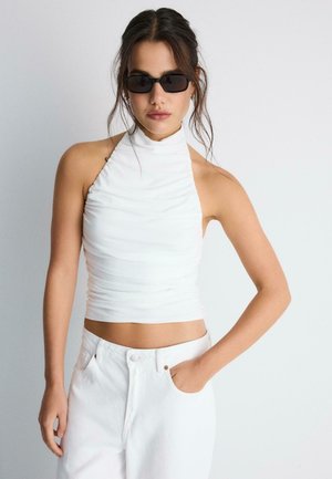 Žena nosiaca čierne slnečné okuliare, biely tielko bez rukávov s vysokým golierom a vysoké biele rifle, jednu ruku v vrecku.