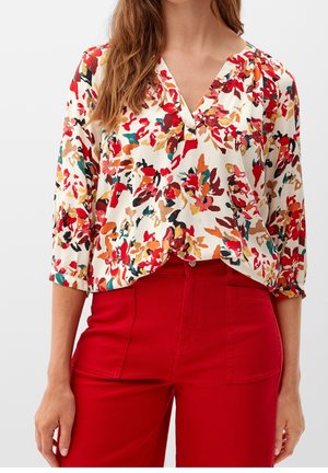 Žena v dlouhém rukávu s květovanou blůzou s červenými, oranžovými a zelenými listy, v kombinaci s vysokým pasem jasně červených kalhot s předními kapsami.