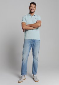 Lichtblauwe polo met een kraag, drie-knoopsluiting, gecombineerd met lichtblauwe jeans en grijze sneakers met witte strepen.