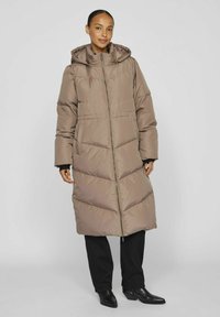 Manteau long en duvet couleur taupe avec une coupe matelassée, fermeture éclair à l'avant et capuche. Dispose de grandes poches latérales et de manches larges. Pantalons et bottes noirs.