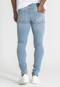 Pohled zezadu na osobu oblečenou v světlých skinny džínách a bílých teniskách, stojící s jednou rukou v zadní kapse na jednobarevném pozadí.