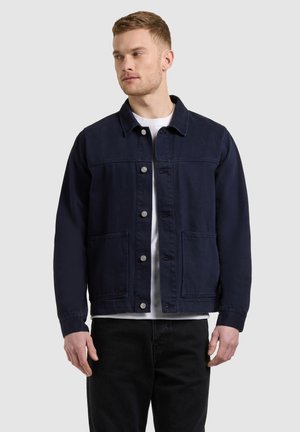 Férfi sötétkék, gombos dzsekiben, fehér inggel és fekete nadrággal, oldalt nézve, egyszínű világos háttér előtt.