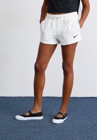 Egy fehér Nike sortot és fekete Mary Jane tornacipőt viselő személy alsóteste kék szőnyegen áll, kezét a zsebébe dugva.
