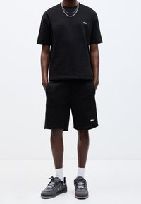 Personne portant une chemise noire à manches courtes, un short noir assorti avec des logos, des chaussettes blanches, des baskets grises et un collier en chaîne argentée sur fond blanc uni.