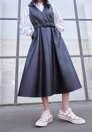 Schwarzes Kunstlederkleid mit tailliertem Oberteil, V-Ausschnitt und weiter Rock, der an der Taille mit einem runden Gürtel gerafft ist; getragen mit weißen Sneakers mit floralen Mustern.