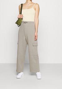 Bežový tílko, šedé široké cargo kalhoty s bočními kapsami a bílé tenisky. Outfit doplňuje malá zelená kabelka na rameno.