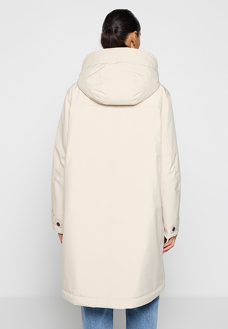 Femme aux cheveux foncés en chignon bas, portant un long manteau beige clair à capuche et un jean bleu, vue de dos.