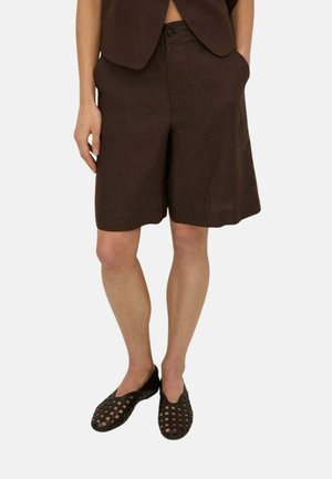 Persona que lleva pantalones cortos de color marrón oscuro hasta la rodilla con bolsillos y zapatos negros tejidos sin cordones, de pie sobre un fondo liso.
