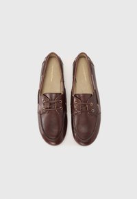 Scarpe da barca in pelle marrone con passanti dorati, punta arrotondata e dettagli cuciti. Suola piatta con una texture liscia e design minimalista.