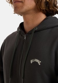 Sort zip-up hoodie med en grøn snor, der har et hvidt logo på brystet. Blødt stof, afslappet pasform og foret hætte.