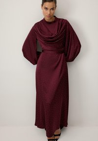 Egy hosszú bordó ruhát viselő nő, apró fehér pöttyökkel, puffos hosszú ujjakkal és lágyan redőzött magas nyakkal, egy fehér fal előtt áll.