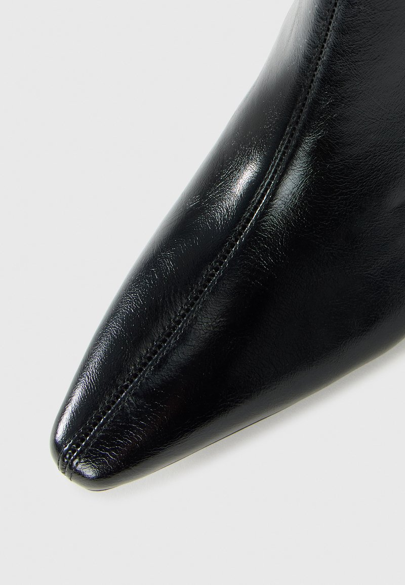 Scarpa di pelle nera lucida con punta affilata e cucitura centrale su uno sfondo chiaro.