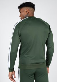 Olivgrüne Sportjacke mit weißen Seitenstreifen, gerippten Bündchen und Saum. Glatter Stoff mit Reißverschlusstaschendetail an der Seite.