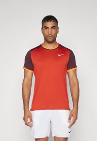 Miestenská športová košeľa v gradientnej červenej farbe, s tmavofialovými rukávmi a oranžovými akcentmi. Vyrobená z priedušnej látky, na hrudi má logo Nike.