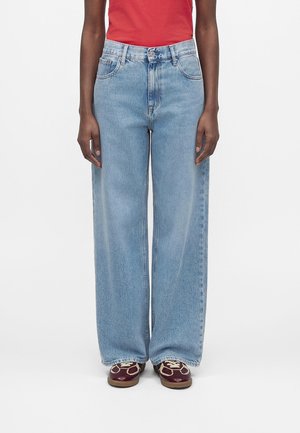 Persoon draagt lichtblauwe wijde spijkerbroek, rood topje en kastanjebruine sneakers met witte veters, staand met ontspannen armen langs het lichaam.