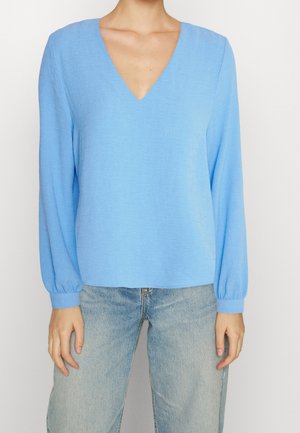 Femme portant un chemisier bleu clair à manches longues et col en V, associé à un jean délavé, debout devant un fond uni.