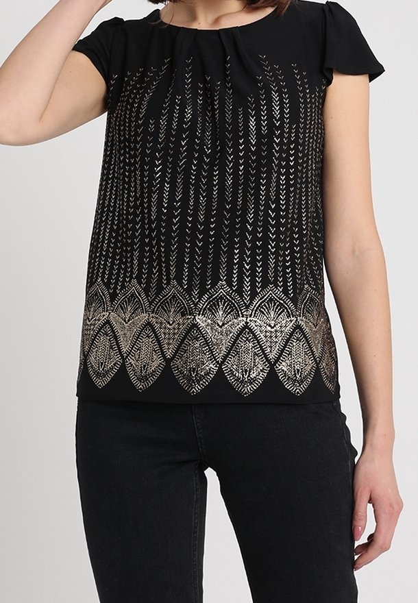 Femme portant un haut noir à manches courtes avec des motifs en forme de flèches et de feuilles argentés métalliques, associé à un pantalon noir.