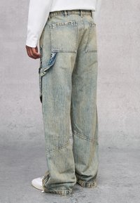 Pantalones cargo de mezclilla sueltos en azul claro, con un acabado desgastado, grandes bolsillos laterales y detalles de costura. Llevados con zapatillas blancas.