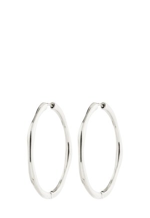 Para srebrzystych, owalnych kolczyków-kołeczek o lekko nieregularnym, falistym kształcie oraz zapięciach na zawiasach na białym tle.