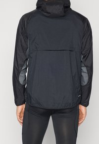 Czarna lekka kurtka z kapturem, z teksturowanej tkaniny, z szarymi bocznymi panelami i tylną kieszenią. Zaprojektowana z myślą o aktywnym ubiorze.