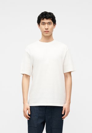 Vīrietis ar īsiem cirtainiem matiem, valkājot vienkāršu balu īss piedurknēm T-kreklu un tumši zilas bikses, stāv pret vienkrāsainu gaišu fonu.