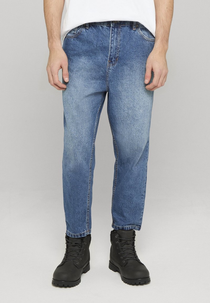 Homme portant un jean bleu délavé et des bottes noires à lacets, debout devant un fond clair uni.
