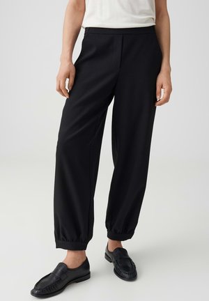 Personne portant un pantalon noir ample et fuselé avec des poignets élastiques, des mocassins noirs et un haut blanc sur un fond clair uni.