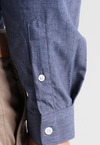 Camisa de manga larga azul oscuro con un tejido texturizado. Presenta un collar puntiagudo, dos botones blancos en los puños y un bolsillo en la manga.