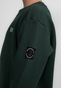 Felpa verde scuro con colletto a girocollo, realizzata in tessuto liscio. Presenta una toppa nera con il logo sulla manica e testo bianco sul petto.