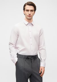 Νεαρός άνδρας που φορεί ανοιχτό ροζ πουκάμισο με κουμπιά, μπουκλωμένο σε γκρι παντελόνι με κορδόνι, στέκεται με το ένα χέρι στην τσέπη σε λευκό φόντο.