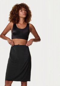 Černá bezešvá podprsenka z hladké látky, doplněná černou sukní ke kolenům s jemným krajkovým lemem. Modelka si upravuje pas sukně.