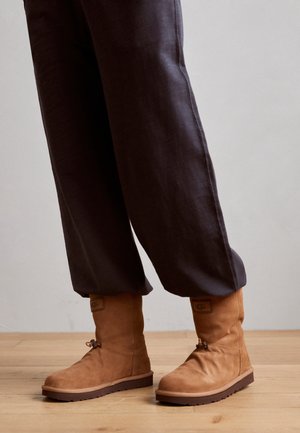 Personne portant un pantalon ample foncé et des bottes en daim couleur fauve mi-mollet avec des semelles en caoutchouc plates, debout sur un sol en bois.