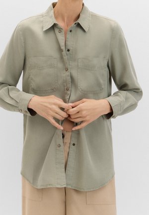Personne boutonnant une chemise manches longues vert olive avec deux poches poitrine, portée avec un pantalon beige sur un fond uni.