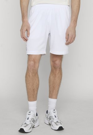 Mann trägt weiße Sportshorts, weiße Kniestrümpfe und weiße New Balance-Sneaker und steht vor einem schlichten Hintergrund.