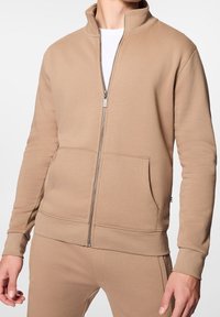 Felpa beige con zip, colletto alto, maniche lunghe, tasche frontali e polsini a coste, realizzata in un morbido tessuto testurizzato.