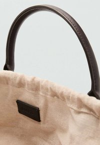 Beige Canvas-Tragetasche mit strukturiertem Finish, ausgestattet mit dunkelbraunen Ledergriffen und einem schwarzen Etikett im Inneren.